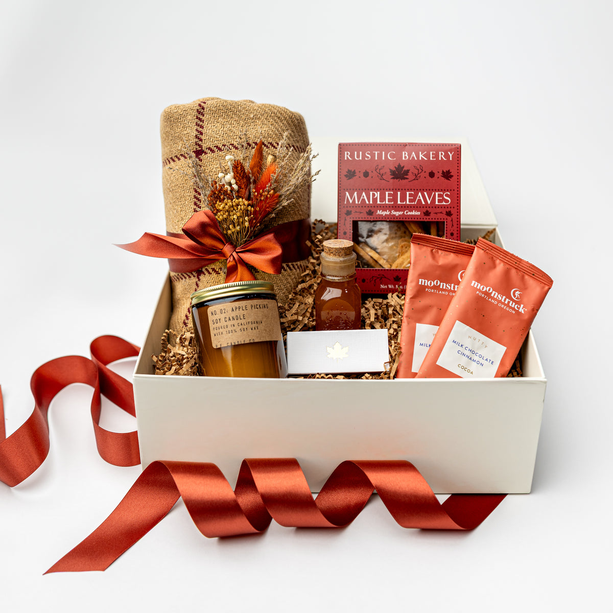 A curated gift box set on a white background, featuring a white box filled with a burlap-wrapped item tied with a rust-red ribbon, a jar candle, a small bottle, a box of Rustic Bakery Maple Leaves tea, and two orange bags from The Artisanal Pantry. A matching rust-red ribbon is draped in front of the box.