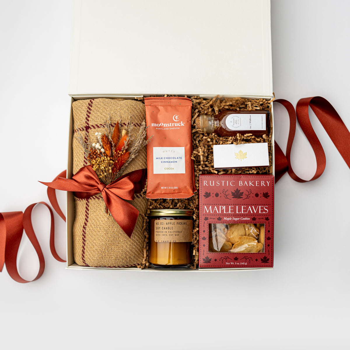 An open white gift box arranged on a white background with autumn-themed items inside: a rolled burlap fabric tied with a rust-orange ribbon, a small orange pouch labeled 'mystruck', a candle in a glass jar, a boxed item labeled '100% CASHmere', and a red box of Rustic Bakery Maple Leaves hand sugar cookies. Decorative dried leaves and a rust-orange ribbon extend from the box.