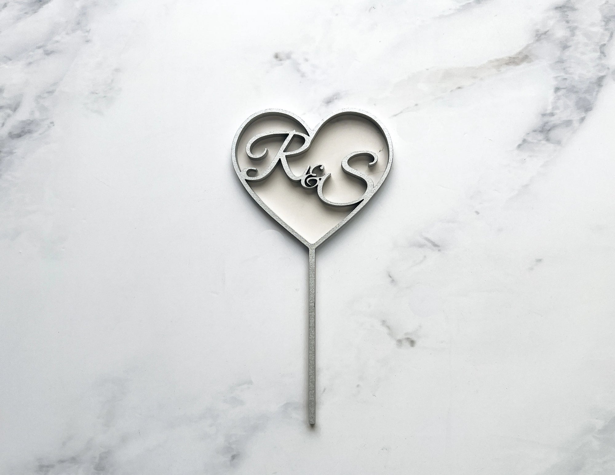 Heart wooden cake topper is featured with the initial R&S in silver in a marble like background.