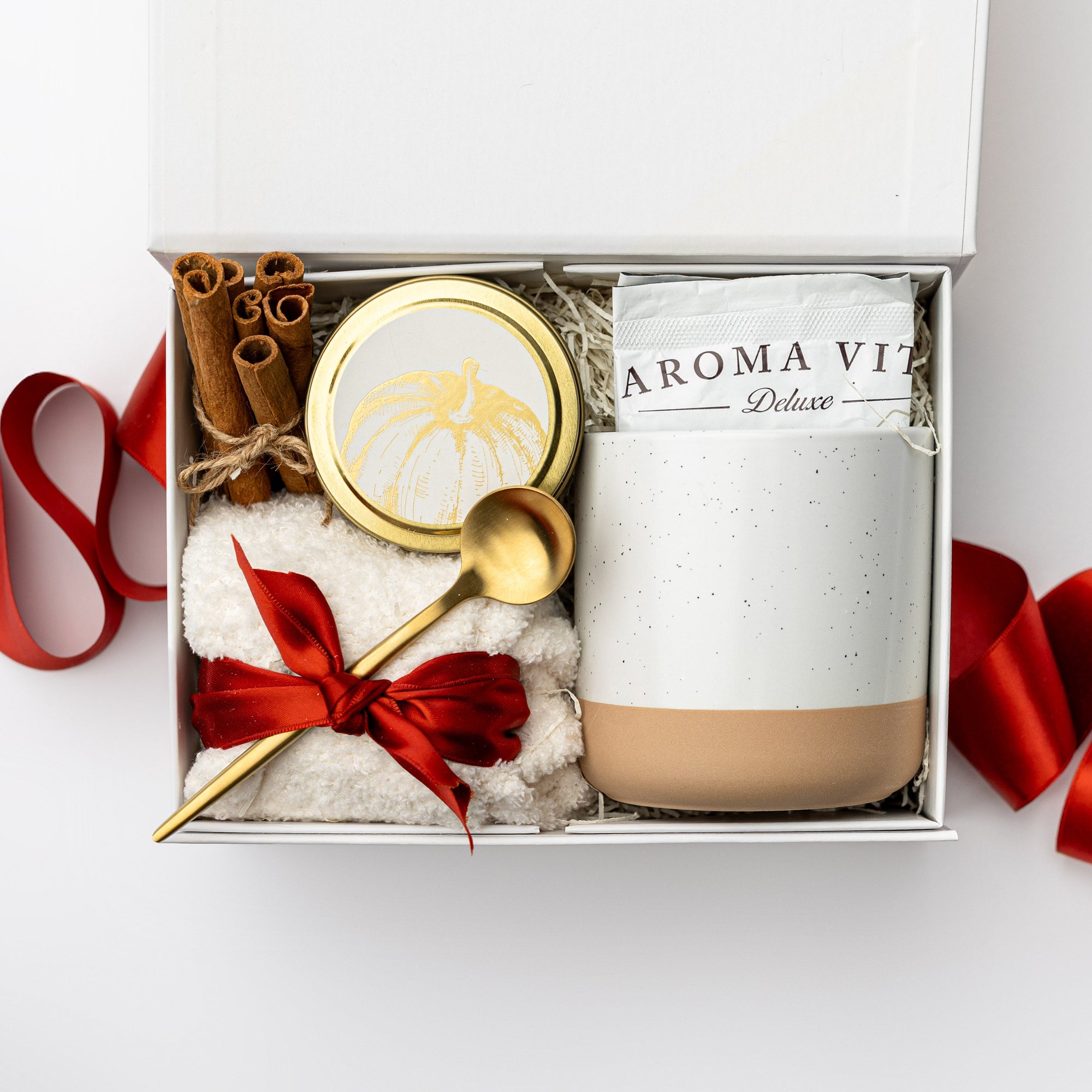 Our Hello Fall gift Box, A top view of our cozy fall gift set featuring a 4 oz. hand-poured Toasted Pumpkin Spice candle, a 10 oz. speckled ceramic mug, two packets of artisan hot cocoa mix, a gold stir spoon, cinnamon sticks, and a pair of unisex ivory cloud socks, all arranged in a white gift box with ivory shred and tied with a red ribbon on a white background.