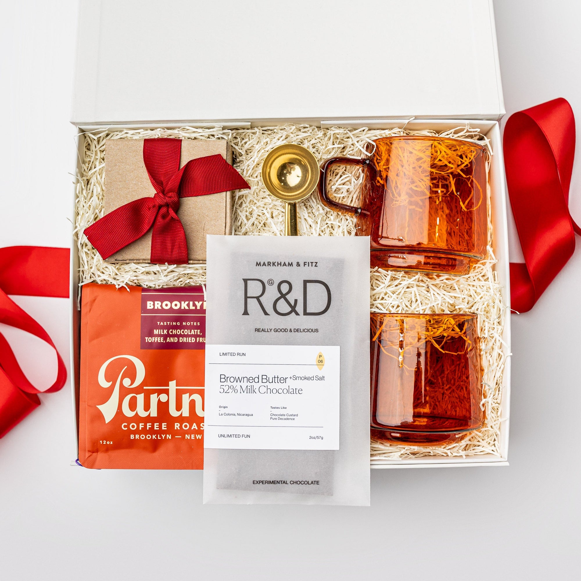 A white gift box on a white background, filled with ivory paper shred. Inside the box are: a 12 oz bag of whole‑bean coffee from Partners (with tasting notes of milk chocolate, toffee, and dried fruit), a set of two modern amber glass mugs, a gold‑metal coffee scoop that also serves as a bag clip, a Markham & Fitz browned butter chocolate bar, and dark‑chocolate sea‑salt caramels — a festive “Holiday Coffee” curated gift box.