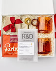 A white gift box on a white background, filled with ivory paper shred. Inside the box are: a 12 oz bag of whole‑bean coffee from Partners (with tasting notes of milk chocolate, toffee, and dried fruit), a set of two modern amber glass mugs, a gold‑metal coffee scoop that also serves as a bag clip, a Markham & Fitz browned butter chocolate bar, and dark‑chocolate sea‑salt caramels — a festive “Holiday Coffee” curated gift box.