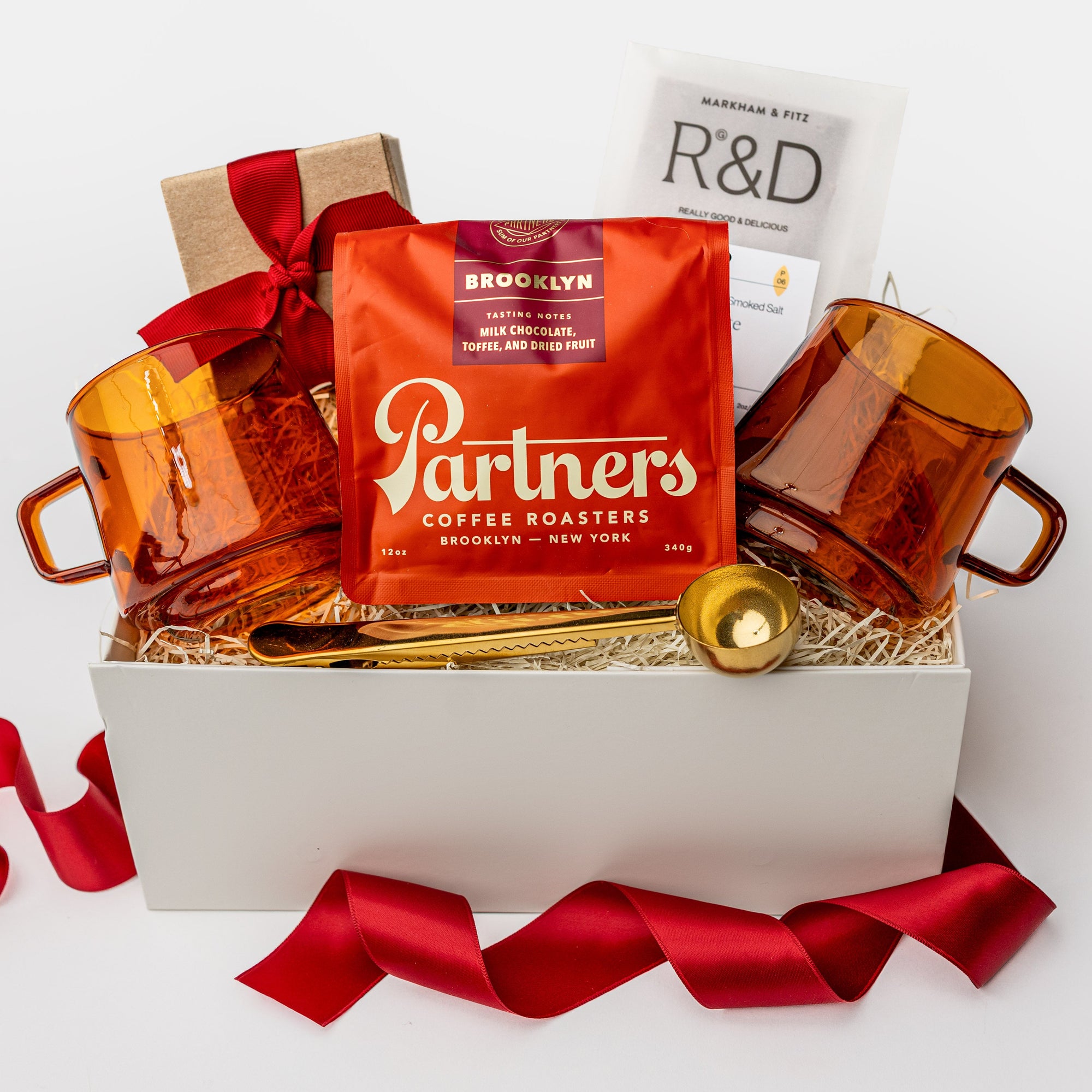 A white gift box on a white background, filled with ivory paper shred. Inside the box are: a 12 oz bag of whole‑bean coffee from Partners (with tasting notes of milk chocolate, toffee, and dried fruit), a set of two modern amber glass mugs, a gold‑metal coffee scoop that also serves as a bag clip, a Markham & Fitz browned butter chocolate bar, and dark‑chocolate sea‑salt caramels — a festive “Holiday Coffee” curated gift box.