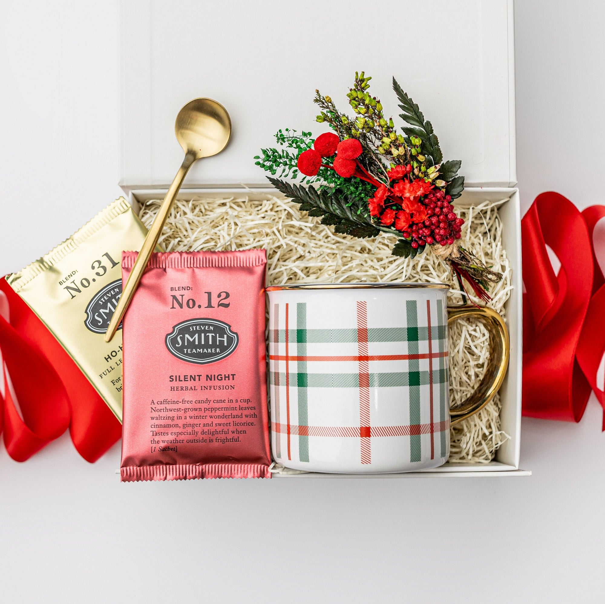 A white gift box with ivory paper shred and tied with a red ribbon, set on a white background. Inside the box: a 12 oz holiday-plaid ceramic mug, two sachets from Smith Teamakers (Ho-Ho-Hojichai and Silent Night), a gold stir spoon, and a handcrafted holiday mini floral bouquet — a curated “Holiday Tea” gift box.