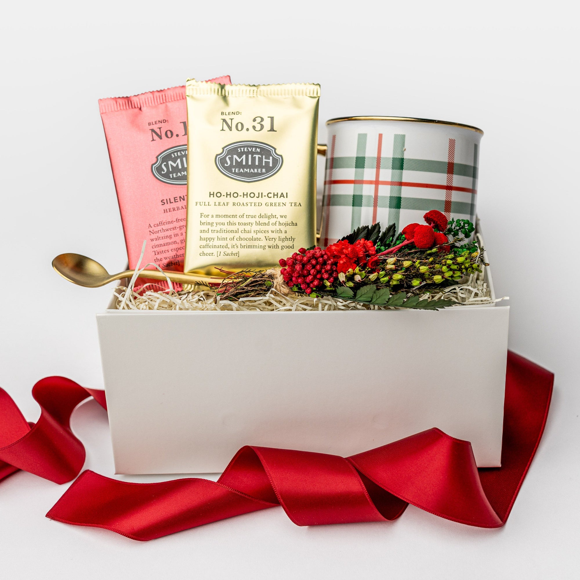 A white gift box with ivory paper shred and tied with a red ribbon, set on a white background. Inside the box: a 12 oz holiday-plaid ceramic mug, two sachets from Smith Teamakers (Ho-Ho-Hojichai and Silent Night), a gold stir spoon, and a handcrafted holiday mini floral bouquet — a curated “Holiday Tea” gift box.