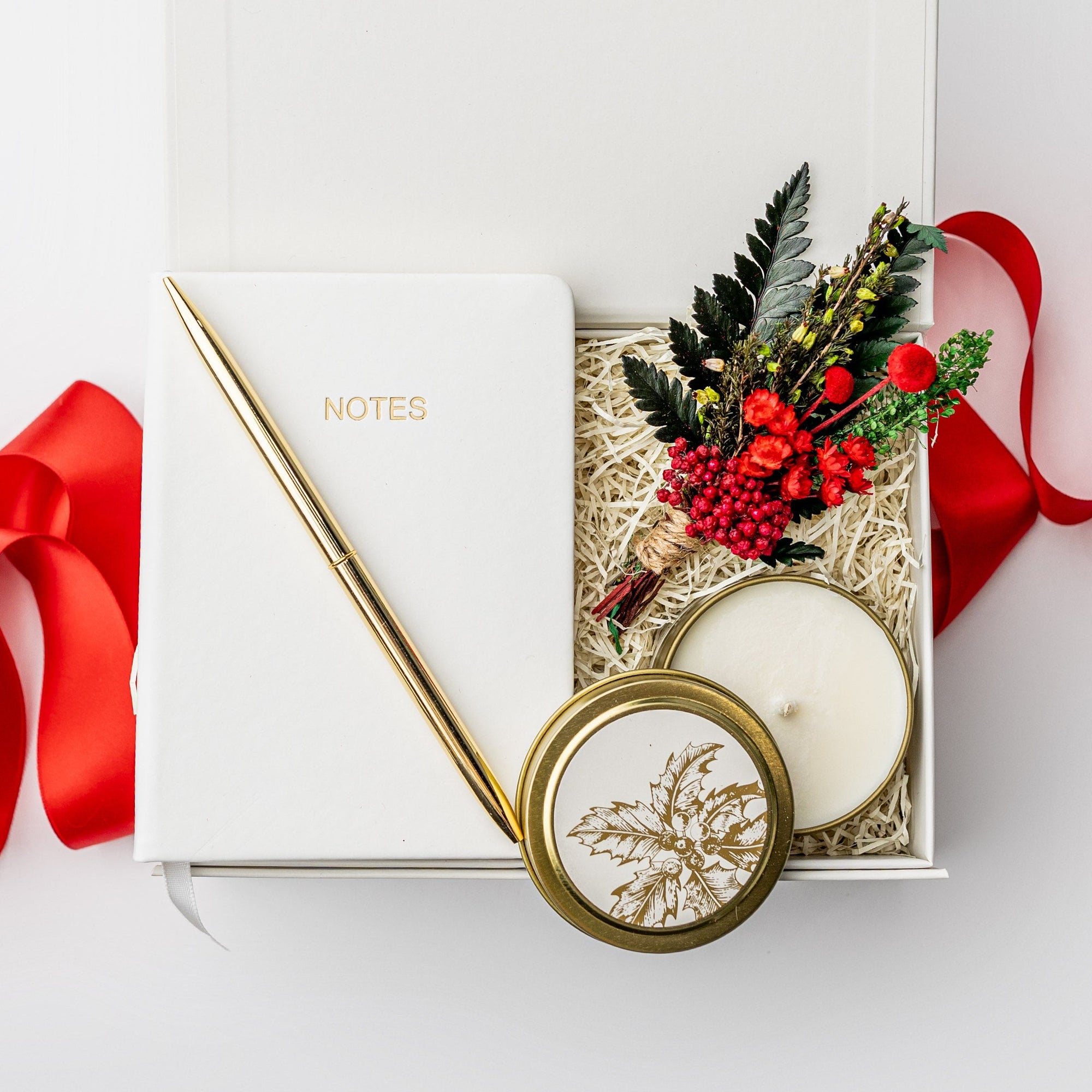 A white gift box with ivory paper shred and tied with a red ribbon on a white background. Inside the box are an ivory soft-cover pocket journal, a minimalist gold bullet pen, a 4oz hand-poured “‘Tis the Season” candle, and a handmade dried floral mini bouquet — a festive “Thankful for You” holiday gift box.