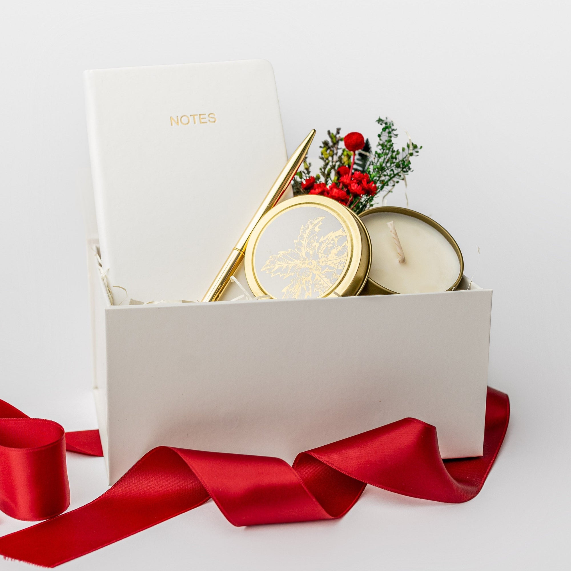 A white gift box with ivory paper shred and tied with a red ribbon on a white background. Inside the box are an ivory soft-cover pocket journal, a minimalist gold bullet pen, a 4oz hand-poured “‘Tis the Season” candle, and a handmade dried floral mini bouquet — a festive “Thankful for You” holiday gift box.