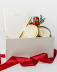 A white gift box with ivory paper shred and tied with a red ribbon on a white background. Inside the box are an ivory soft-cover pocket journal, a minimalist gold bullet pen, a 4oz hand-poured “‘Tis the Season” candle, and a handmade dried floral mini bouquet — a festive “Thankful for You” holiday gift box.