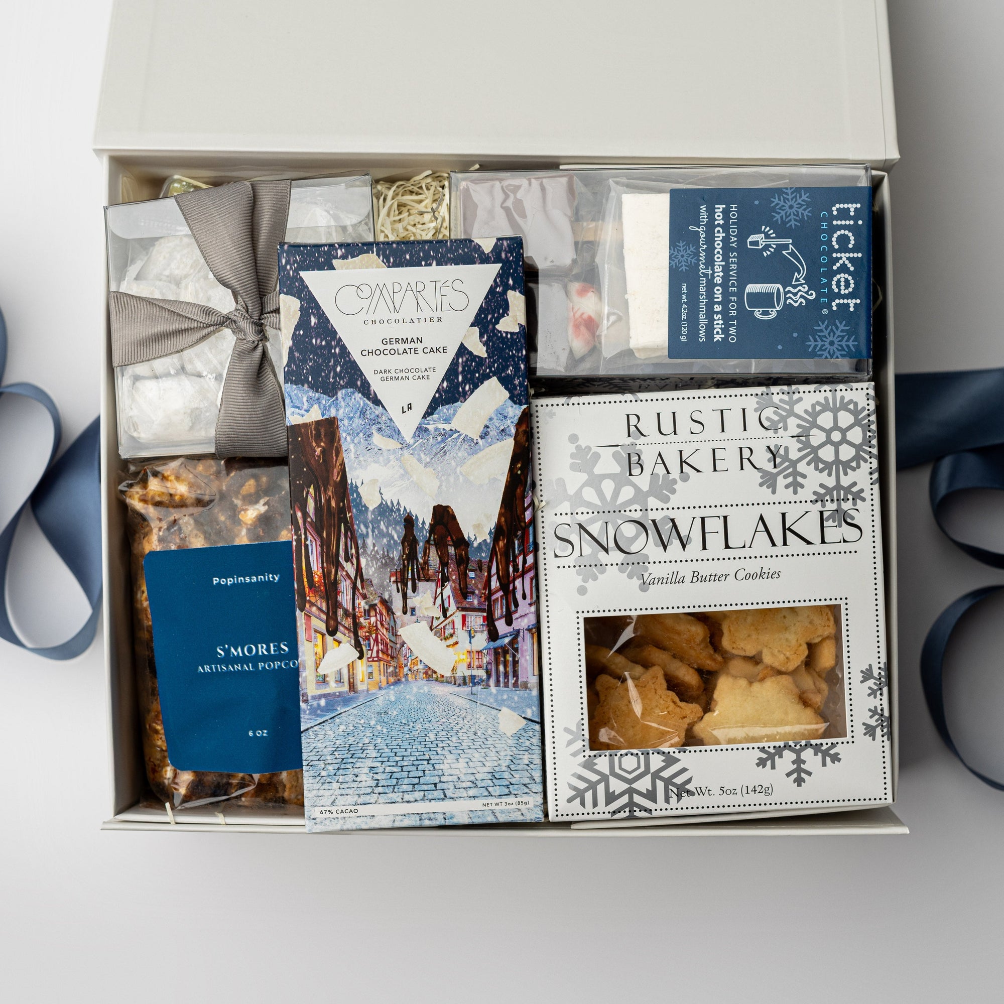 Holiday Treats Gift Box is featured with a blue ribbon crossing underneath. Contents include a German Chocolate Cake Bar, Tea Cakes (11.2 oz), S’mores Popcorn (6 oz), Snowflakes Vanilla Butter Cookies (5 oz), a 2-pack of Hot Cocoa on a Stick, Almond Toffee Treats (7.94 oz), and Sea Salt Caramel Candy (8 oz), all arranged on ivory shred. The image is displayed on a white background.