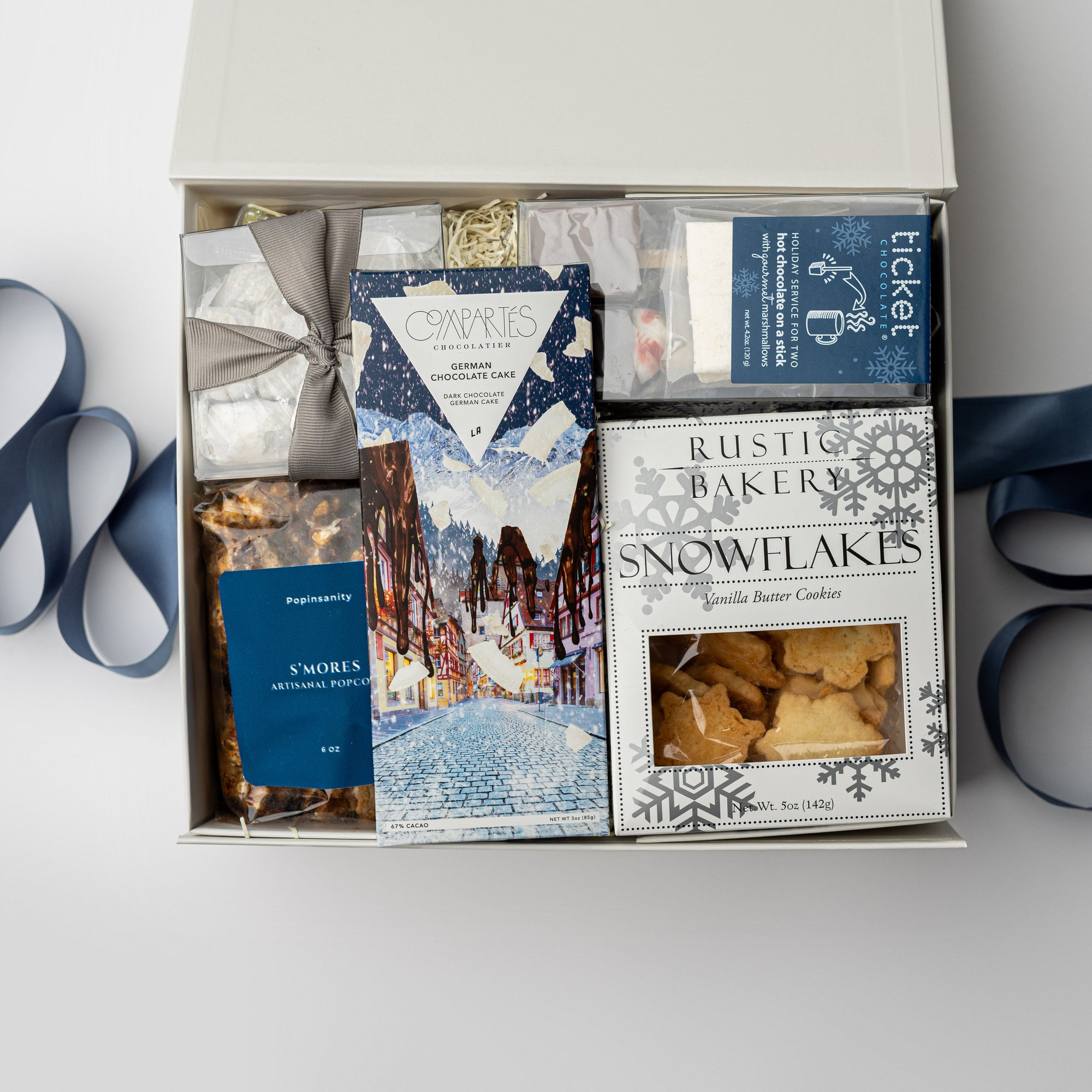 Holiday Treats Gift Box is featured with a blue ribbon crossing underneath. Contents include a German Chocolate Cake Bar, Tea Cakes (11.2 oz), S’mores Popcorn (6 oz), Snowflakes Vanilla Butter Cookies (5 oz), a 2-pack of Hot Cocoa on a Stick, Almond Toffee Treats (7.94 oz), and Sea Salt Caramel Candy (8 oz), all arranged on ivory shred. The image is displayed on a white background.