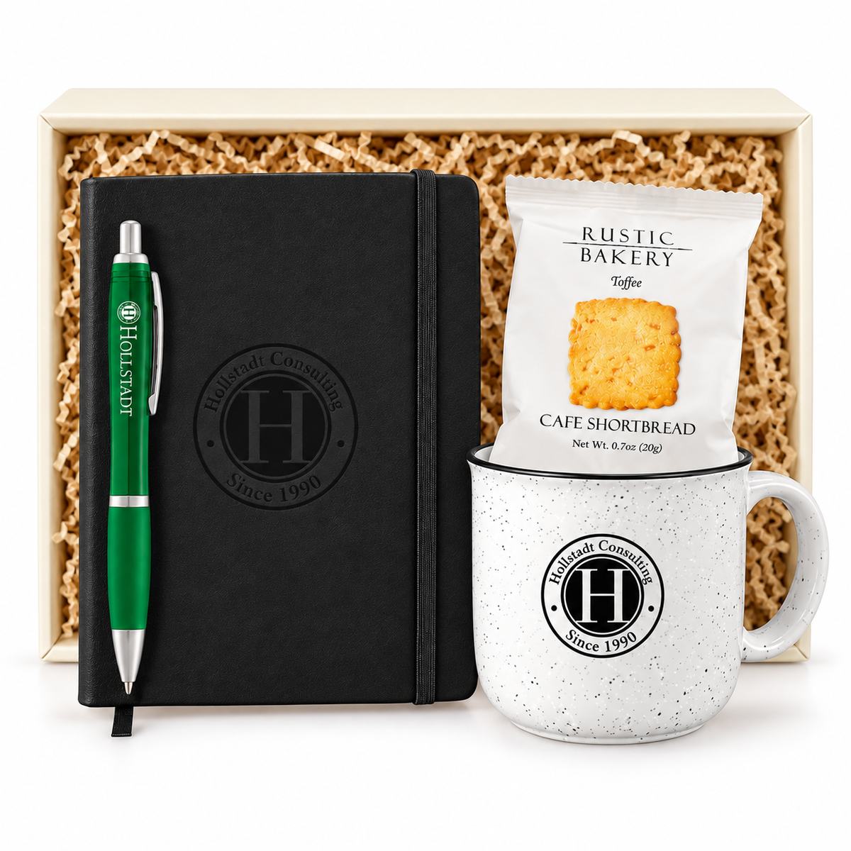A gift set featuring a black notebook with a green pen, a white ceramic mug, and a bag of Rustic Bakery Toffee Cafe Shortbread cookies, all arranged in front of a wooden box filled with crinkle paper.