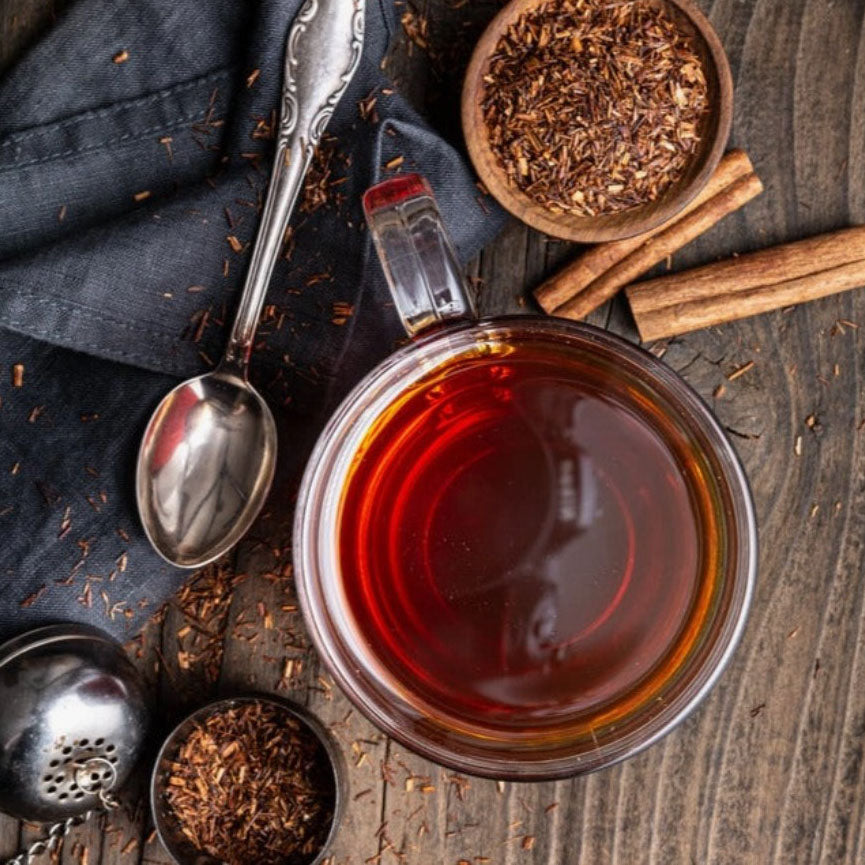 A high-angle close-up view of a cup of reddish-brown tea, accompanied by loose tea, a spoon, and cinnamon sticks on a dark wooden surface.

The image  features a centrally placed clear glass cup filled with tea.  The cup is positioned in the center of the image. The dark wooden surface provides a rustic and natural backdrop,  Loose tea is scattered around the cup, and a metal tea infuser bottom left, a dark grey cloth draped with tea behind, silver spoon, bowl with tea and cinnamon sticks around. 