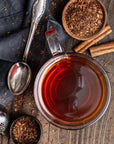 A high-angle close-up view of a cup of reddish-brown tea, accompanied by loose tea, a spoon, and cinnamon sticks on a dark wooden surface.

The image  features a centrally placed clear glass cup filled with tea.  The cup is positioned in the center of the image. The dark wooden surface provides a rustic and natural backdrop,  Loose tea is scattered around the cup, and a metal tea infuser bottom left, a dark grey cloth draped with tea behind, silver spoon, bowl with tea and cinnamon sticks around. 