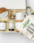 A top view of the New Home Housewarming Gift Box featuring a set of two marble and wood coasters, a “Home Sweet Home” tea towel, a 9 oz. hand-poured candle labeled “New Beginnings, New Home, New Memories” (optionally personalized with a family name), a mini tea tube of jasmine green tea, a mini jar of honey with a wooden honey drizzler, and a set of two speckled stoneware mugs, all arranged in a white gift box with ivory shred and tied with a green ribbon on a white background.