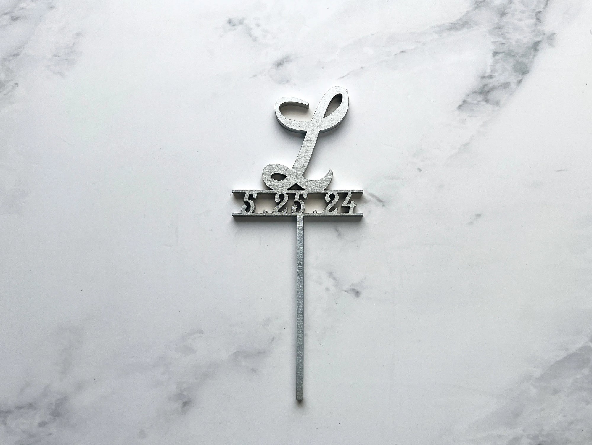 Initial & date wooden cake topper is featured personalized with L and 5.325.24 in silver on a marble like background.