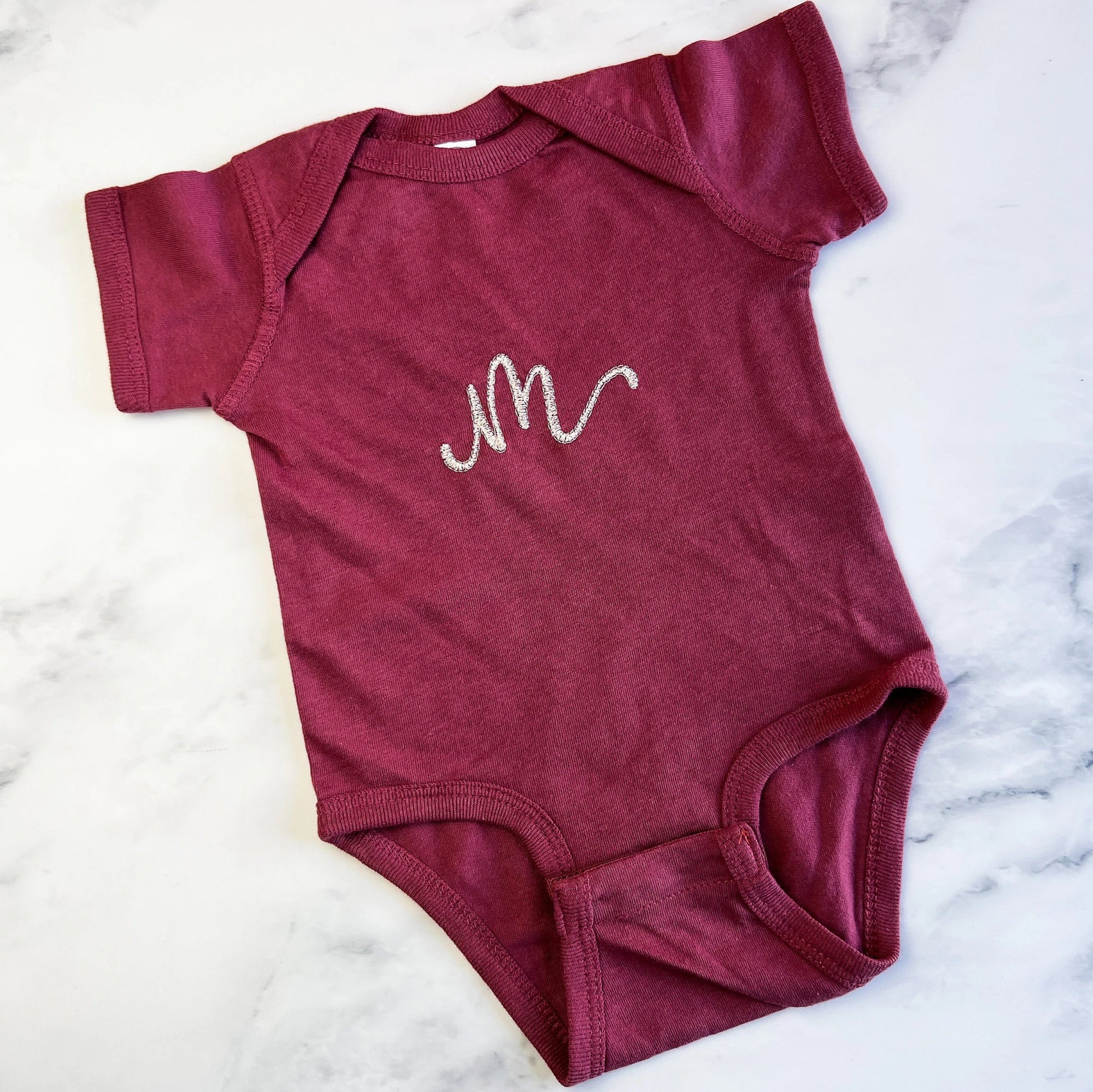 Initial embroidered onesie in rouge personalized in champagne thread with the letter M on a marble like background. 