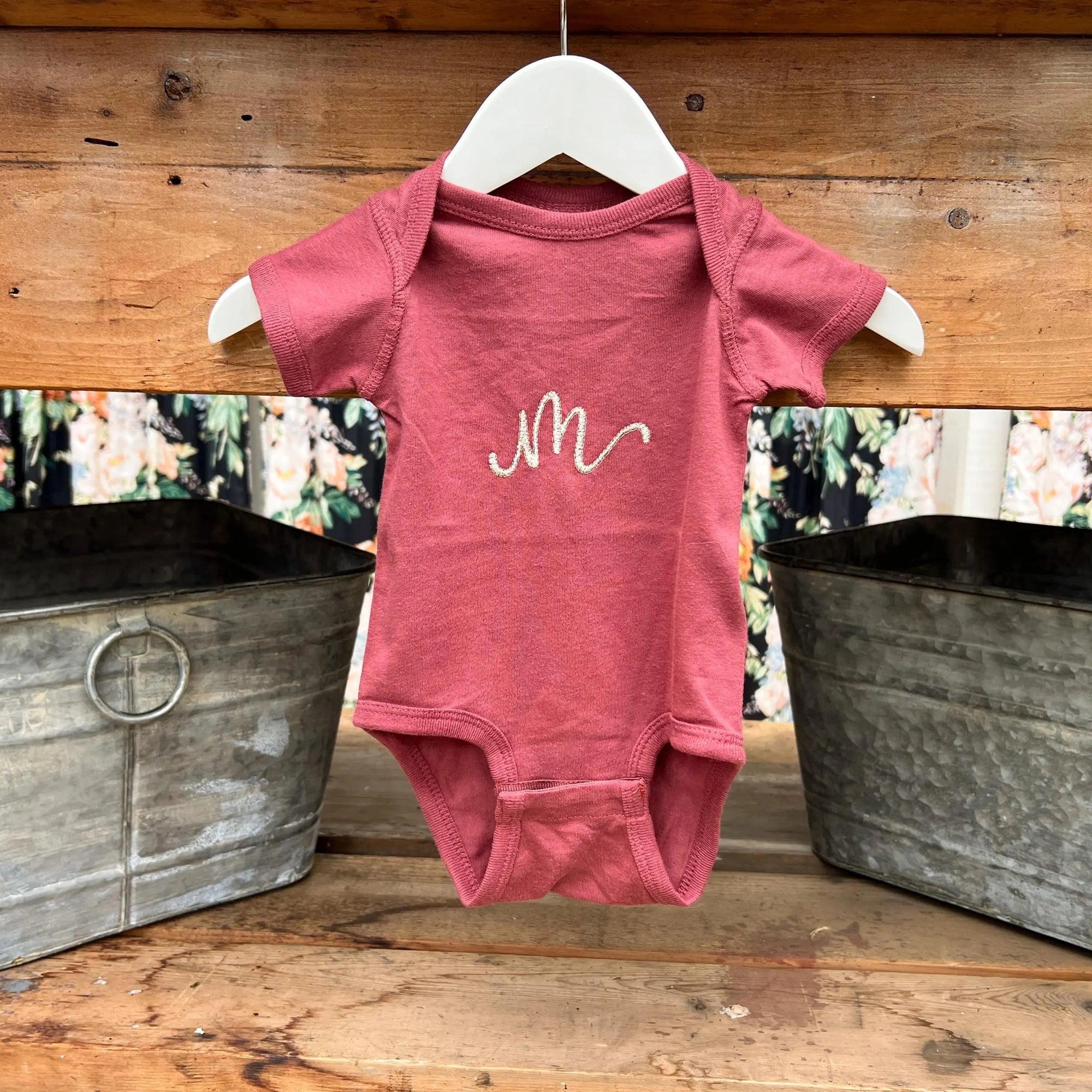 Initial embroidered onesie in rouge personalized in champagne thread with the letter M hanged in a white hanger in a wooden background with two silver buckets on each side. 