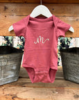 Initial embroidered onesie in rouge personalized in champagne thread with the letter M hanged in a white hanger in a wooden background with two silver buckets on each side. 