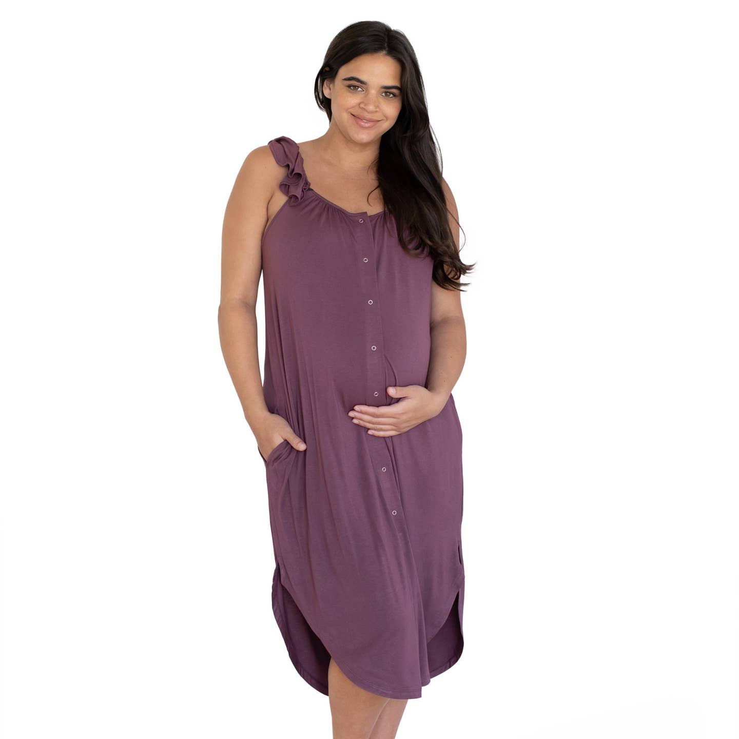Kindred bravely ruffle strap & delivery gown the image displays a woman with long, dark hair parted to the side, framing her face. She has fair skin and a gentle smile, with what appear to be freckles on her face, and her eyes show a relaxed, contented expression. She is wearing a sleeveless, button-down dress in a muted purple color, made of soft-looking fabric, that drapes comfortably around her body. The dress features a ruffled detail on the shoulder straps.  