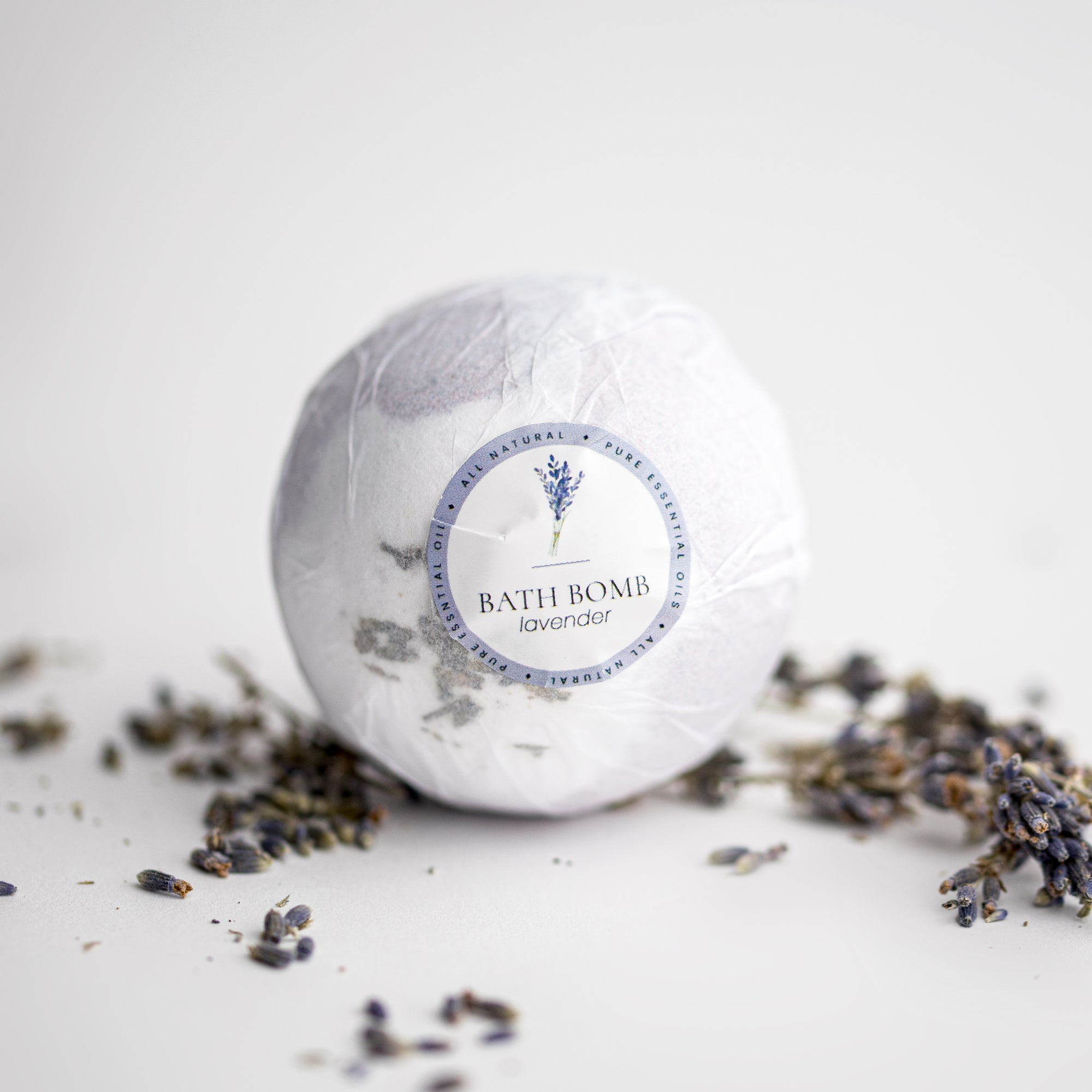 Lavender bath bomb, round-shaped, features in its white transparent wrapping with a white label with purple details, a lavender image, and black lettering. Surrounded by lavender pieces on a white background. 