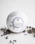 Lavender bath bomb, round-shaped, features in its white transparent wrapping with a white label with purple details, a lavender image, and black lettering. Surrounded by lavender pieces on a white background. 