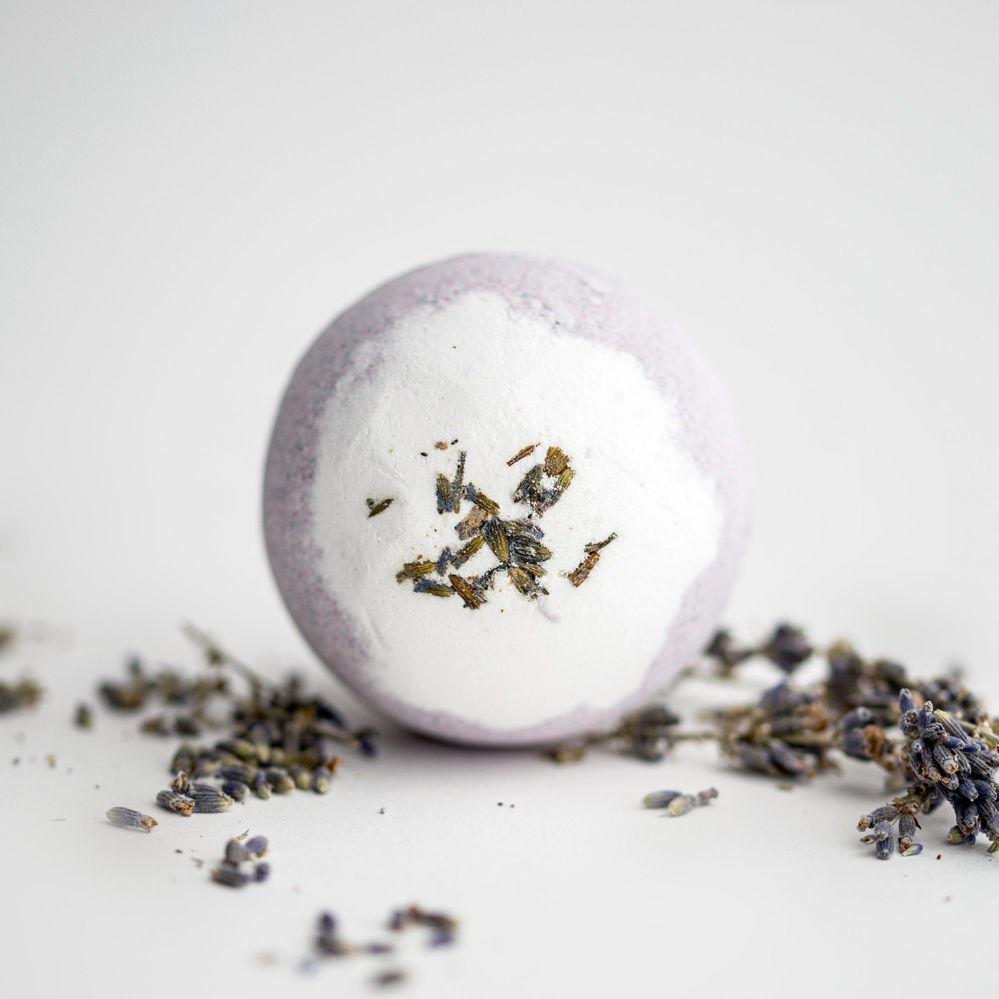 Lavender bath bomb, round-shaped, surrounded by lavender pieces on a white background. 