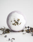Lavender bath bomb, round-shaped, surrounded by lavender pieces on a white background. 