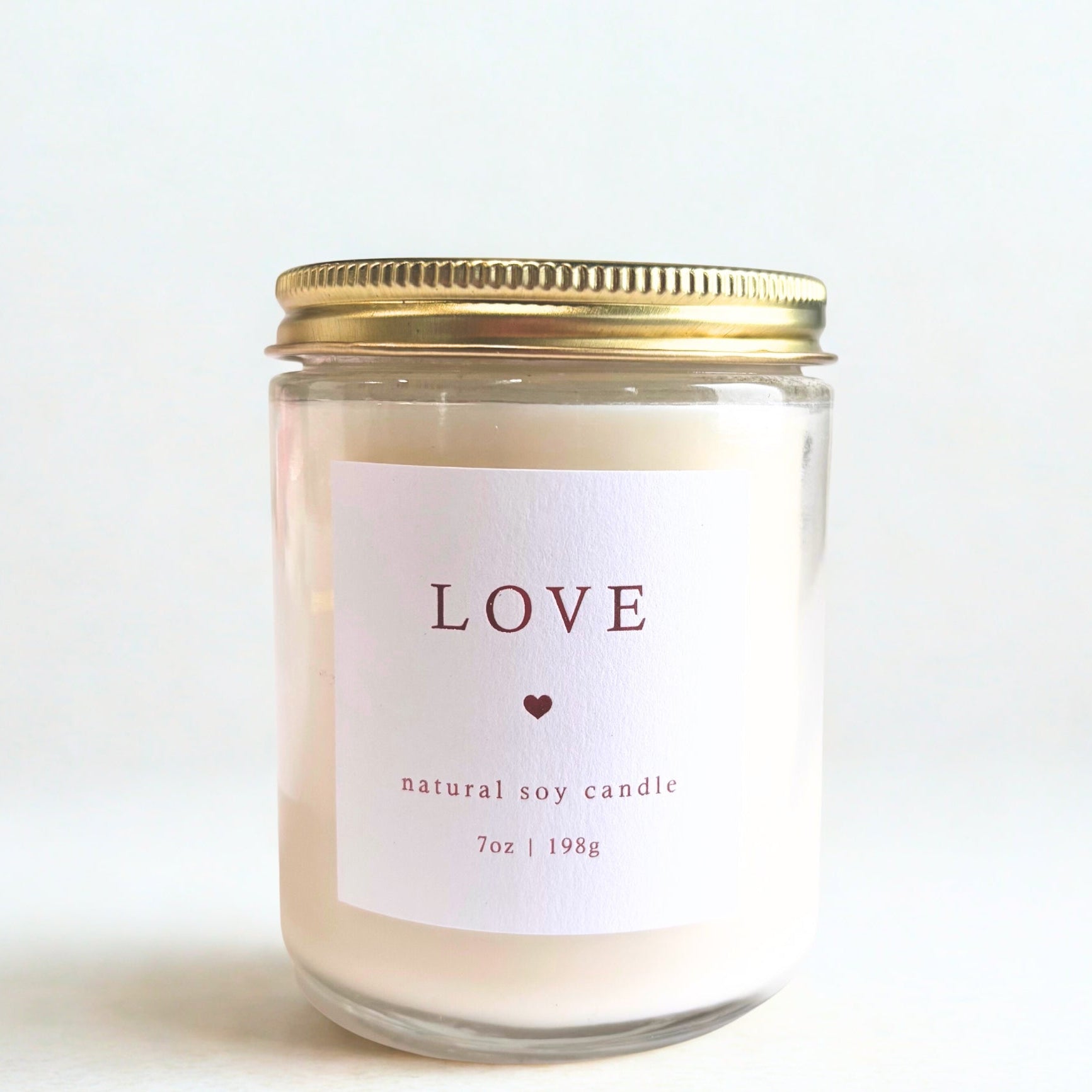 Love candle is featured in a transparent jar with gold lid with a white label wit red lettering.
