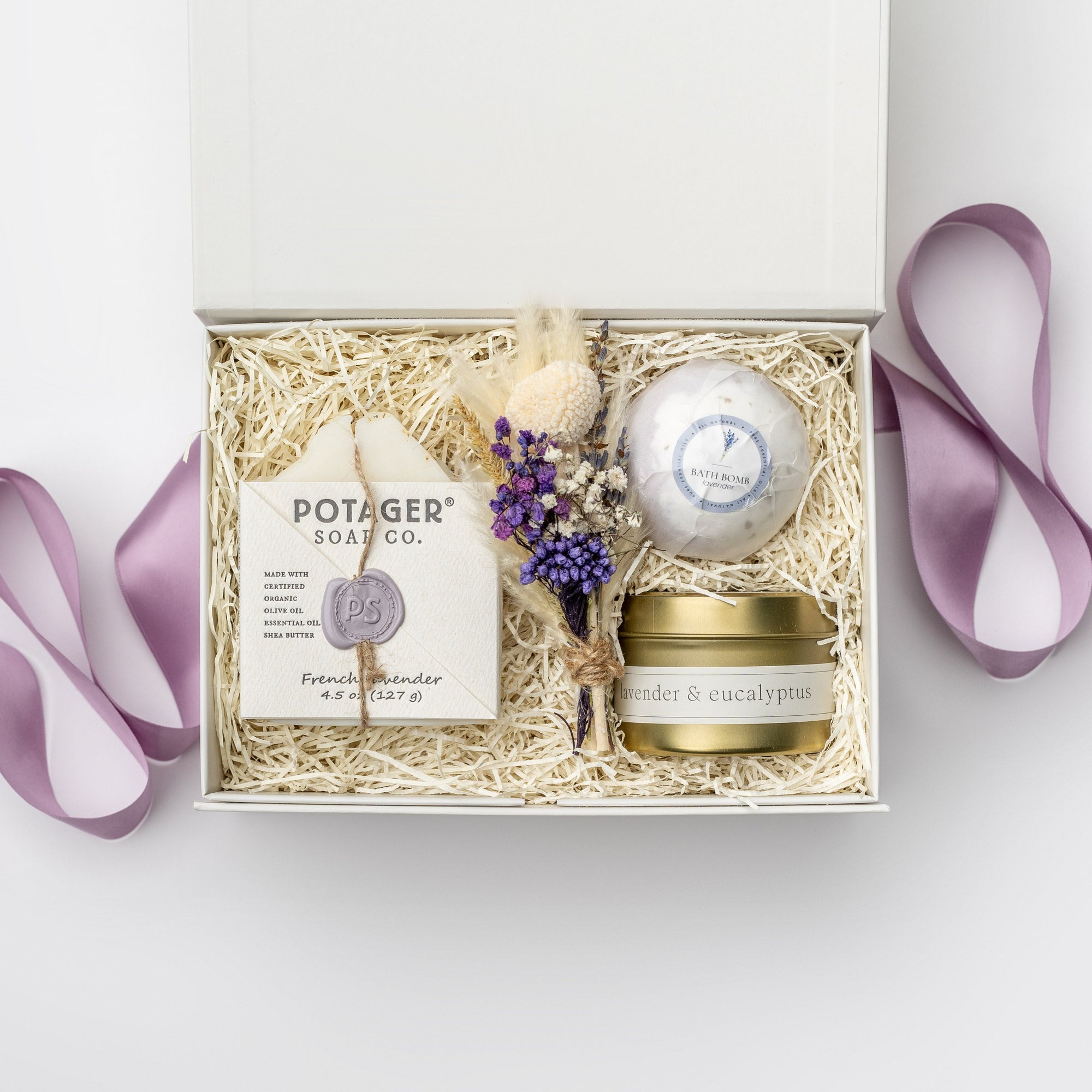 A top view of our lovely lavender gift box, a lavender-themed gift set featuring a lavender bath bomb, a 4 oz. hand-poured lavender candle, French lavender soap, and a handmade dried floral mini bouquet, all arranged in a white gift box with ivory shred and tied with a purple ribbon on a white background