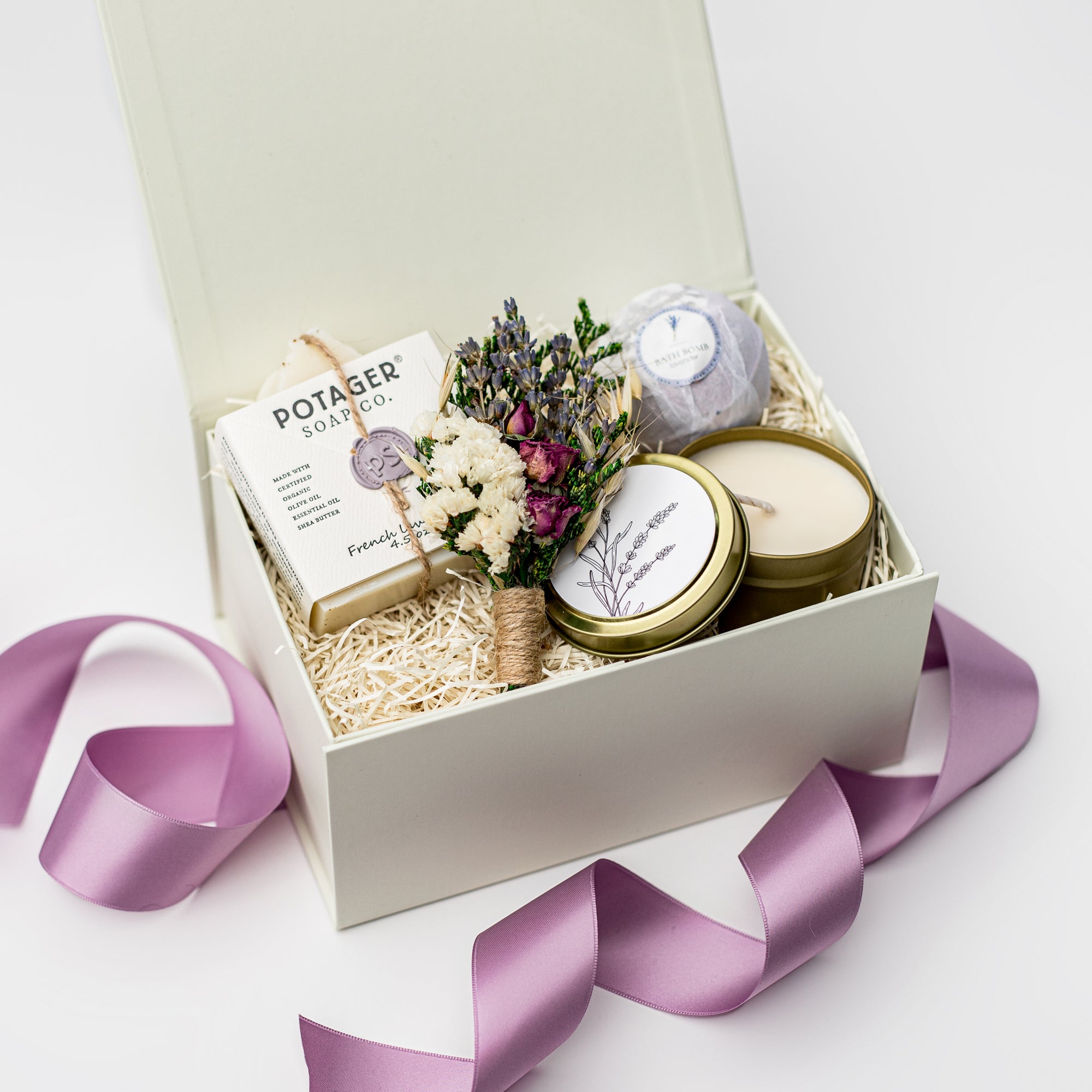 A top view of a lavender-themed gift set featuring a lavender bath bomb, a 4 oz. hand-poured lavender candle, French lavender soap, and a handmade dried floral mini bouquet, all arranged in a white gift box with ivory shred and tied with a purple ribbon on a white background.