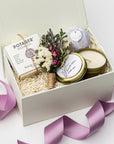 A top view of a lavender-themed gift set featuring a lavender bath bomb, a 4 oz. hand-poured lavender candle, French lavender soap, and a handmade dried floral mini bouquet, all arranged in a white gift box with ivory shred and tied with a purple ribbon on a white background.