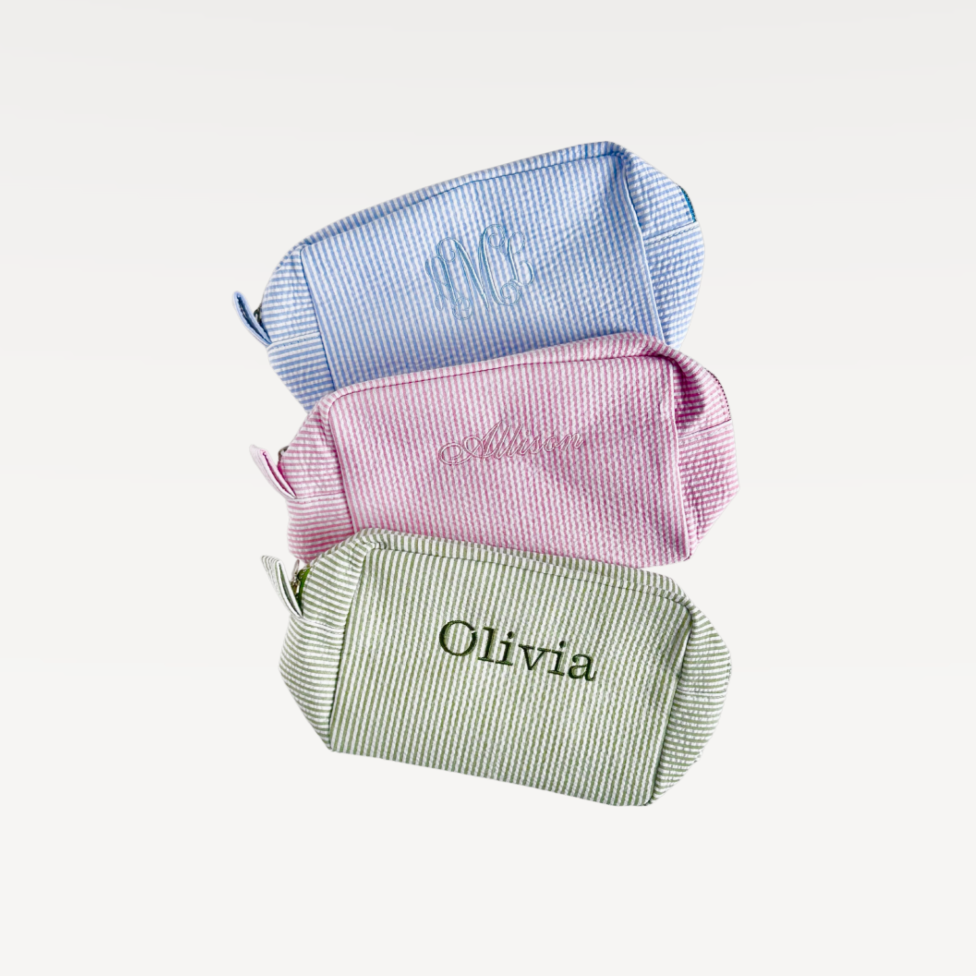 Three small fabric pouches in blue, pink, and green with embroidered names on a light gray background.