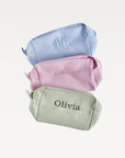 Three small fabric pouches in blue, pink, and green with embroidered names on a light gray background.