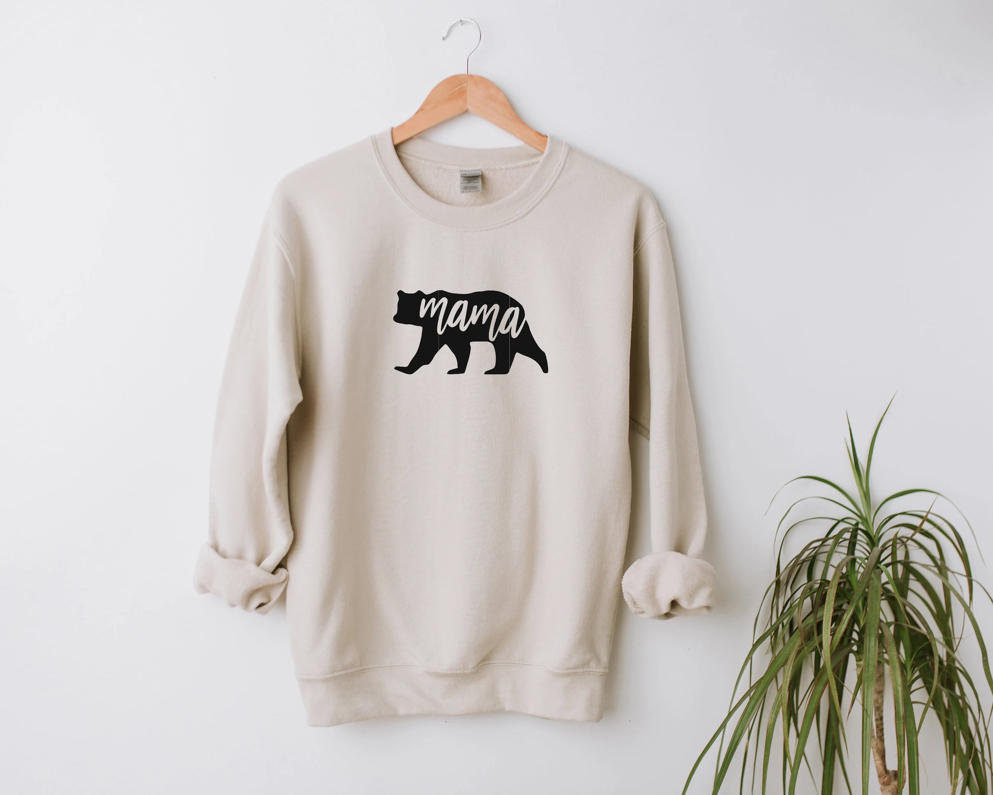 Mama bear sweatshirt featured in sand color engraved with bear silhouette with mama cut out on the middle of the image hanged in a white background with a plant in the bottom right corner. 