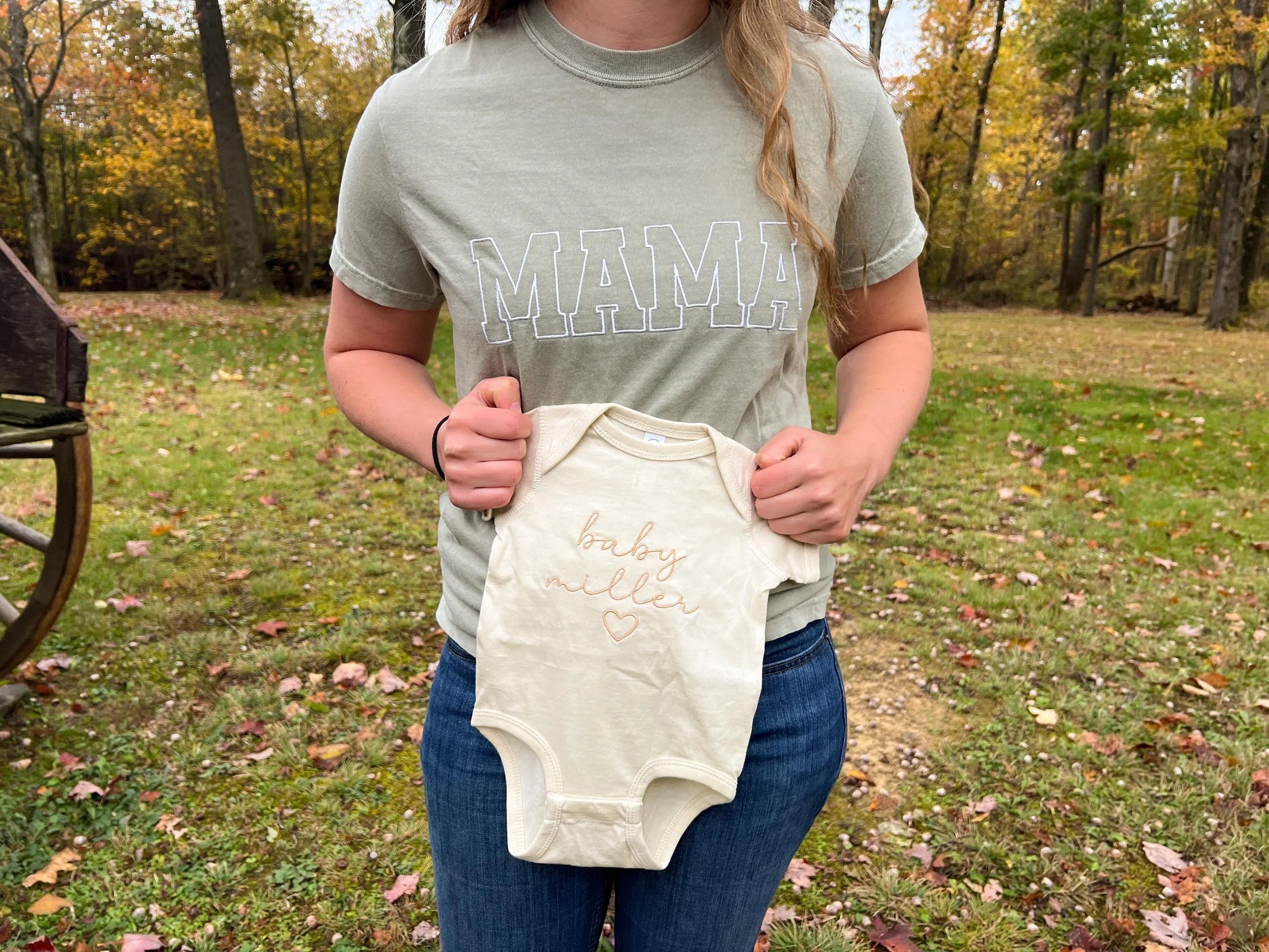 A mother wearing the embroidered comfort color tee in bay color with mama embroidered in white thread. on a wood nature background holding a onesie personalized baby Miller.