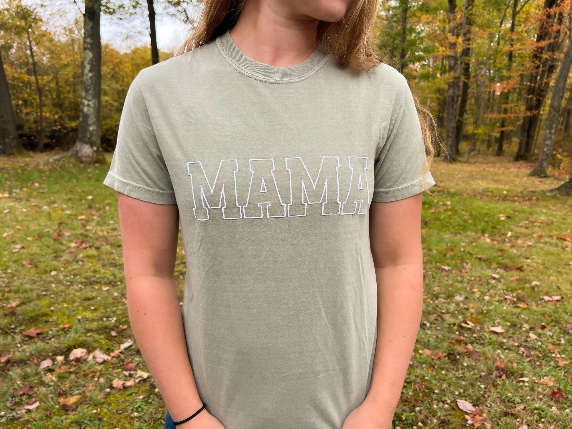 A mother wearing the embroidered comfort color tee in bay color with mama embroidered in white thread. on a wood nature background.