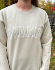 Woman wearing an ivory sweatshirt with the word ‘MAMA’ in white letters across the chest, standing outdoors in front of a rustic wagon and autumn foliage.