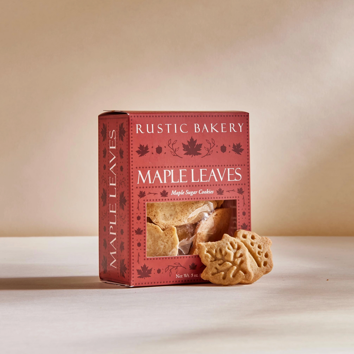 A box of Rustic Bakery Maple Leaves baked cookies sits on a light beige surface against a warm beige background. Two leaf-shaped cookies are placed in front of the box, and a clear window on the front of the red packaging shows the cookies inside.