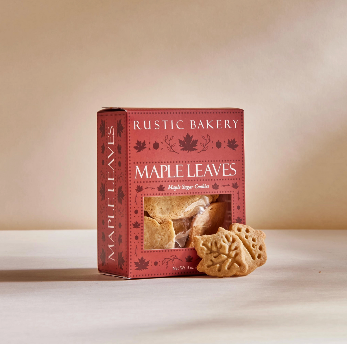 A box of Rustic Bakery Maple Leaves baked cookies sits on a light beige surface against a warm beige background. Two leaf-shaped cookies are placed in front of the box, and a clear window on the front of the red packaging shows the cookies inside.