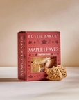 A box of Rustic Bakery Maple Leaves baked cookies sits on a light beige surface against a warm beige background. Two leaf-shaped cookies are placed in front of the box, and a clear window on the front of the red packaging shows the cookies inside.