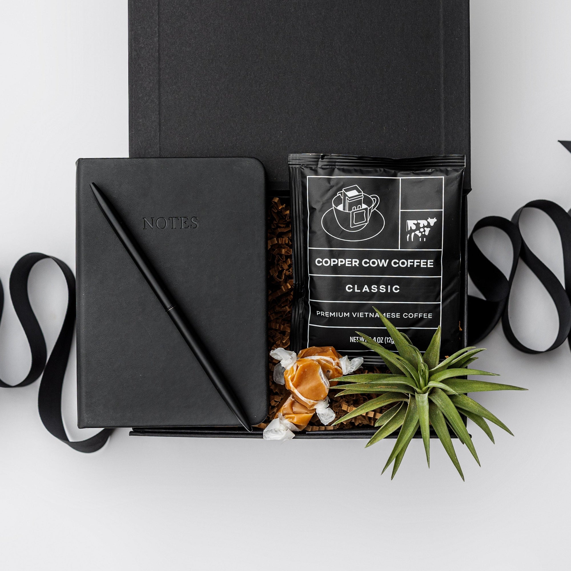 Top view of our Merci Gift Box, presented in a black gift box with brown shredded paper and tied with a black ribbon, featuring a black vegan leather pocket notebook, a matte black minimalist pen, an artisan single-serve pour-over coffee, taffy caramels, and a small air plant.