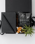 Top view of our Merci Gift Box, presented in a black gift box with brown shredded paper and tied with a black ribbon, featuring a black vegan leather pocket notebook, a matte black minimalist pen, an artisan single-serve pour-over coffee, taffy caramels, and a small air plant.