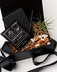 Top view of our Merci Gift Box, presented in a black gift box with brown shredded paper and tied with a black ribbon, featuring a black vegan leather pocket notebook, a matte black minimalist pen, an artisan single-serve pour-over coffee, taffy caramels, and a small air plant.