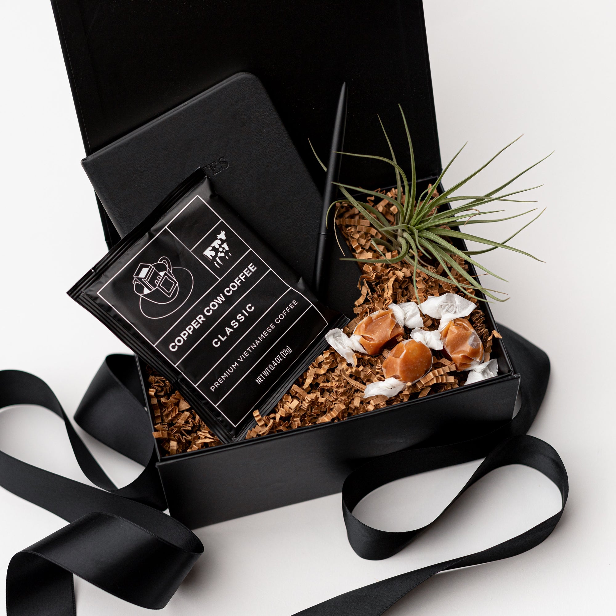 Top view of our Merci Gift Box, presented in a black gift box with brown shredded paper and tied with a black ribbon, featuring a black vegan leather pocket notebook, a matte black minimalist pen, an artisan single-serve pour-over coffee, taffy caramels, and a small air plant.