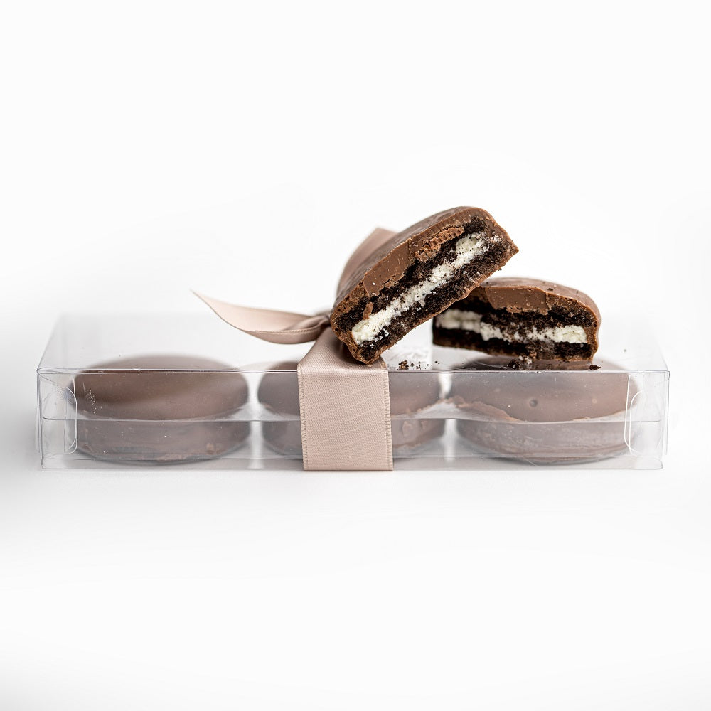 Milk chocolate-covered Oreos featured in a transparent rectangular package tied with a beige ribbon, with three inside and two placed on top of the package.