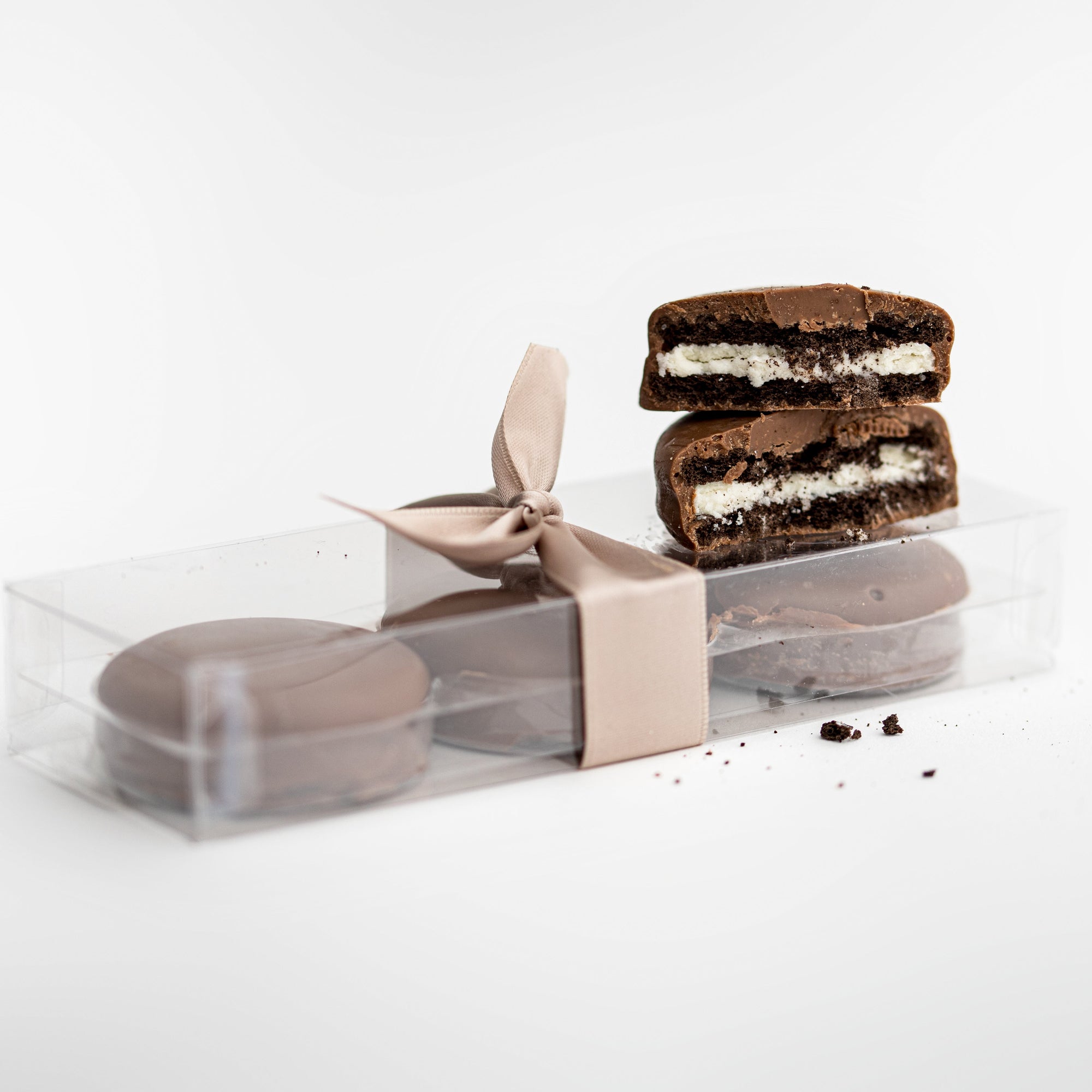 Milk chocolate-covered Oreos, vertical view, featured in a transparent rectangular package tied with a beige ribbon, with three inside and two placed on top of the package.
