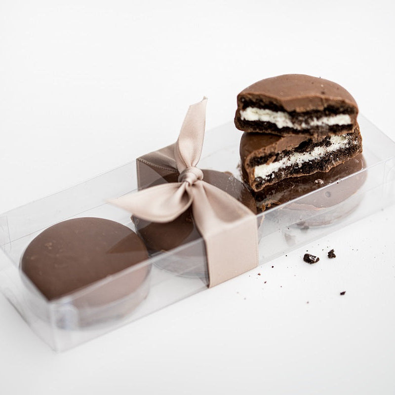 Milk chocolate-covered Oreos, vertical view, featured in a transparent rectangular package tied with a beige ribbon, with three inside and two placed on top of the package.