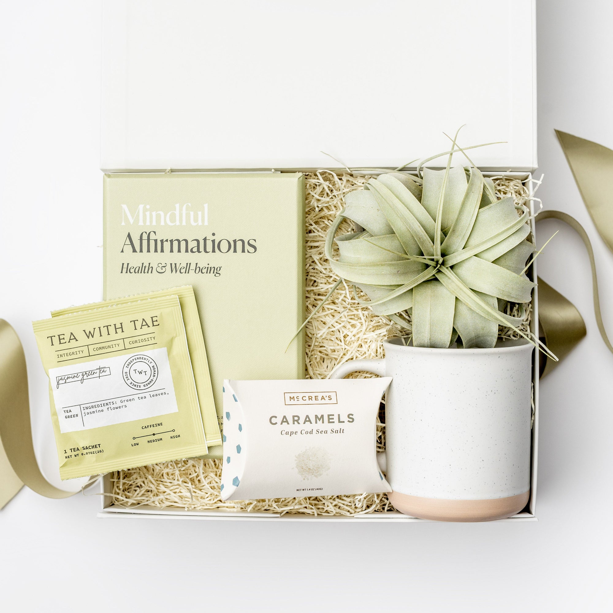 A top view of the Mindful Meditation Gift Box featuring a set of Mindful Affirmation Cards for Health & Wellbeing, a Tillandsia Xerographica live air plant, two green jasmine tea sachets, a 12 oz. speckled ceramic mug, and handcrafted Cape Cod sea salt caramels, all arranged in a white gift box with ivory shred and tied with a green ribbon on a white background.