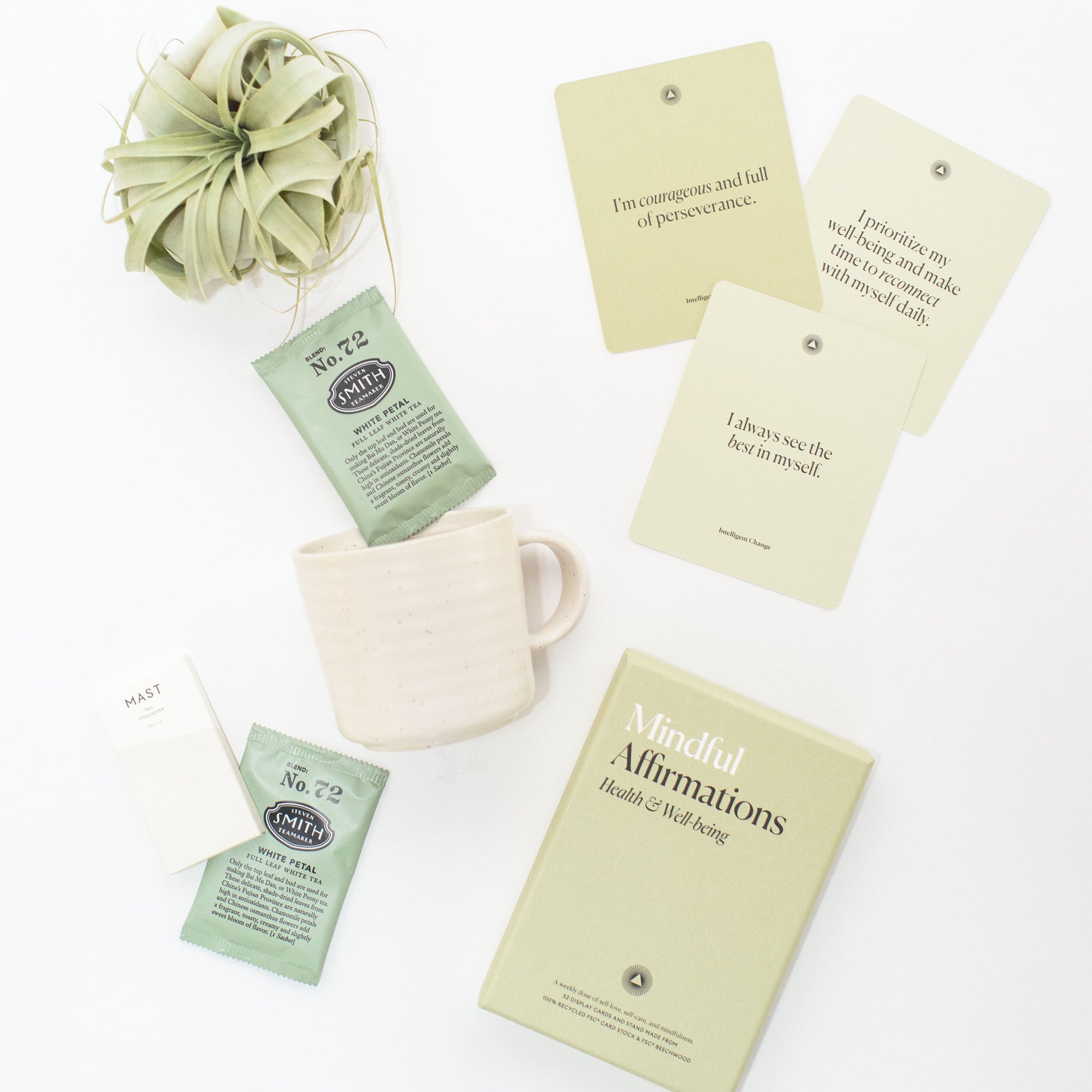 Meditation Gift Box items displayed on a white background, featuring Mindful Affirmation Cards, relaxing tea, a stoneware mug, an artisan chocolate bar, and a green air plant.