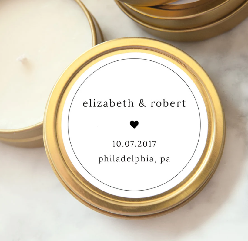 Close up image of minimal heart custom wedding favor candle shown in its gold tin with. a white round label with Elizabeth & Robert a heart the date 10.07.2017 philadelphia pa location 